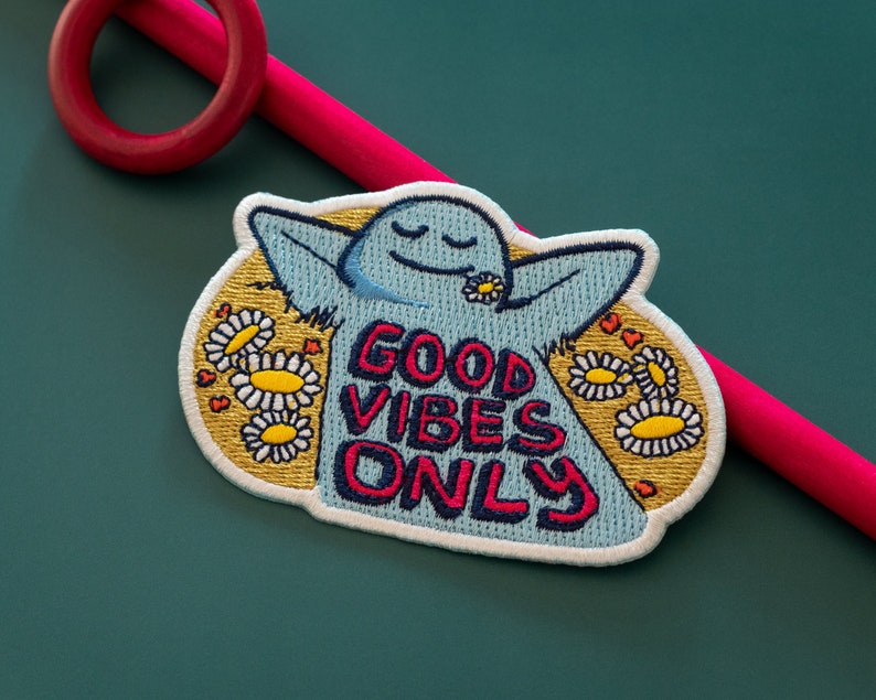 K&ouml;nnte beinhalten: Ein blauer gestickter Aufn&auml;her mit einer Comicfigur mit geschlossenen Augen und einer Blume im Mund. Die Figur ist von gelben G&auml;nsebl&uuml;mchen umgeben und der Text "GOOD VIBES ONLY" ist in Pink und Blau geschrieben.