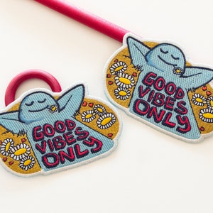 K&ouml;nnte beinhalten: Zwei blaue gestickte Aufn&auml;her mit einer Comicfigur und dem Text "Good Vibes Only", umgeben von gelben G&auml;nsebl&uuml;mchen und roten Herzen.