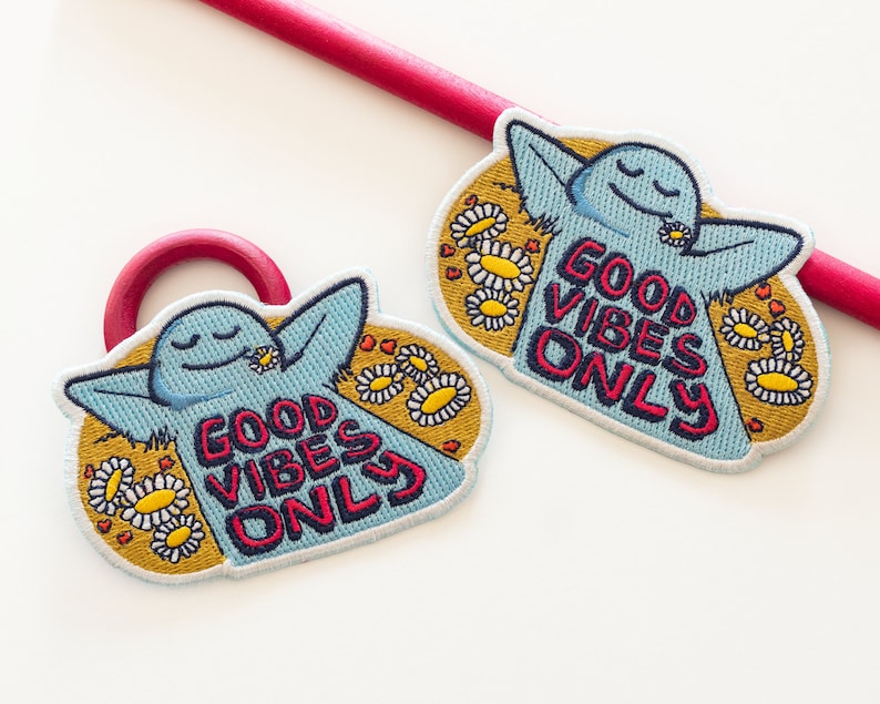 K&ouml;nnte beinhalten: Zwei blaue gestickte Aufn&auml;her mit einer Comicfigur und dem Text "Good Vibes Only", umgeben von gelben G&auml;nsebl&uuml;mchen und roten Herzen.