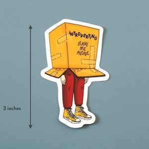 May include: A yellow and brown cardboard box with the text "Introverting Leave Me Alone" on the front. A person with red pants and yellow shoes is wearing the box as a hat. The sticker is 3 inches tall.