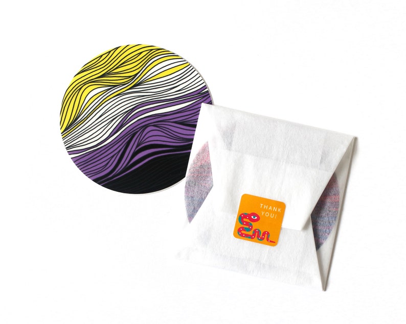 May include: A round coaster with a purple, white, and yellow striped design. The coaster is on a white surface with a small orange square sticker that says "Thank You!" with a red snake illustration.