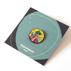 May include: A colorful enamel pin featuring a cluster of pink, green, and blue mushrooms on a yellow and orange background. The pin is circular and has a black metal backing. The text "RippleSplash Studio" is printed on the card.
