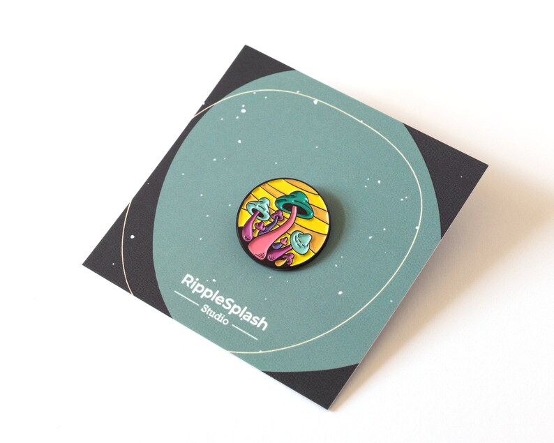 May include: A colorful enamel pin featuring a cluster of pink, green, and blue mushrooms on a yellow and orange background. The pin is circular and has a black metal backing. The text "RippleSplash Studio" is printed on the card.