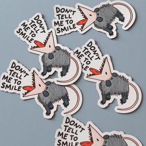 May include: Five stickers featuring a cartoon opossum with the text "Don't tell me to smile". The opossum is gray with a pink mouth and a red tongue.