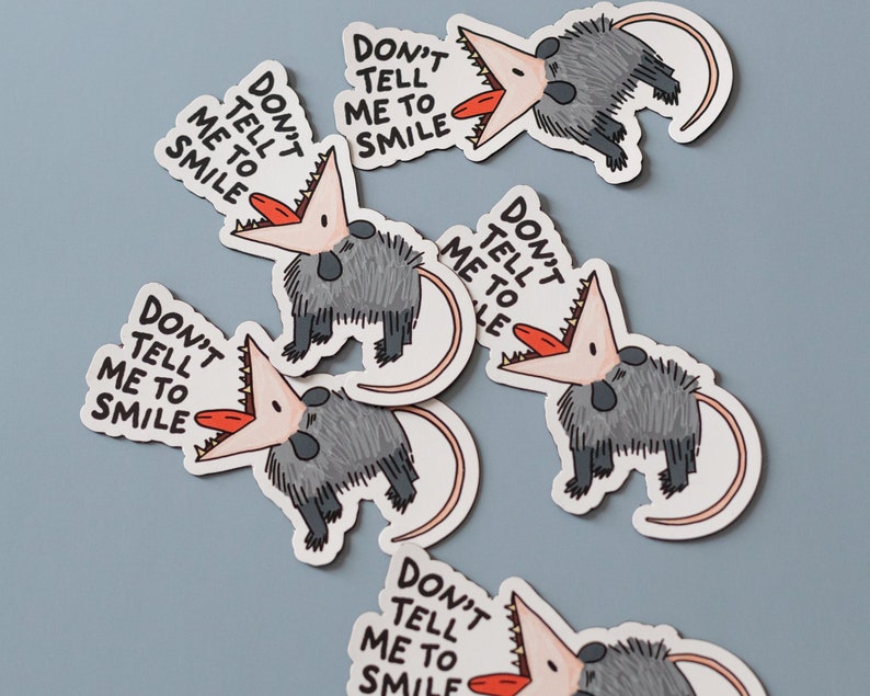 May include: Five stickers featuring a cartoon opossum with the text "Don't tell me to smile". The opossum is gray with a pink mouth and a red tongue.