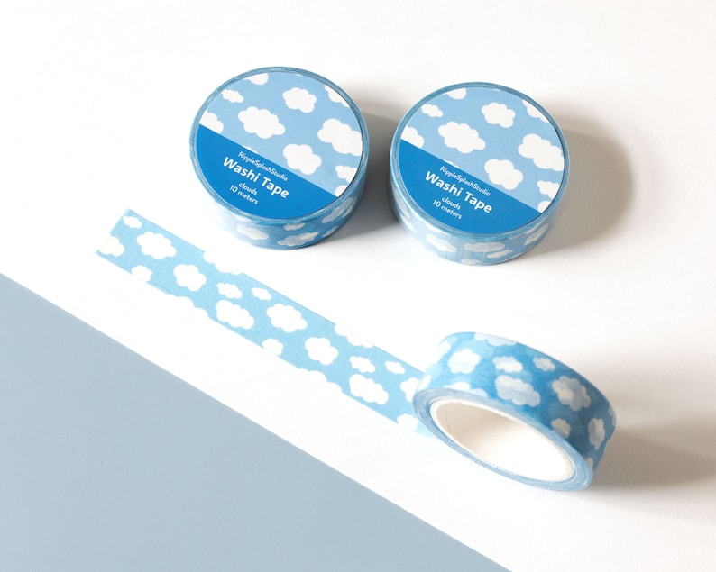 May include: Two rolls of blue washi tape with a white cloud pattern, and a strip of tape unrolled. The tape rolls have the text "Washi Tape" and "clouds 10 meters". The background is white and light blue.