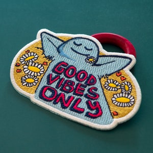 K&ouml;nnte beinhalten: Ein blauer gestickter Aufn&auml;her mit einem l&auml;chelnden Gesicht und dem Text "Good Vibes Only", umgeben von gelben Blumen. Der Aufn&auml;her hat eine rote Schlaufe zum Anbringen.