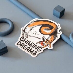 May include: A white sticker with a cartoon corgi chasing its tail, with the text "CHASING DREAMS" in black. The corgi is orange, brown, and white. The sticker is on a light blue surface with blue geometric shapes.