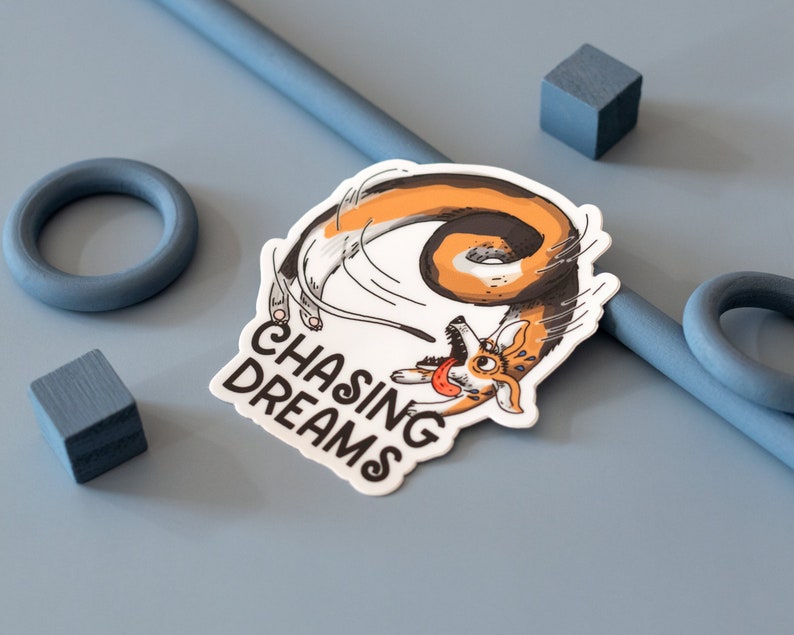 May include: A white sticker with a cartoon corgi chasing its tail, with the text "CHASING DREAMS" in black. The corgi is orange, brown, and white. The sticker is on a light blue surface with blue geometric shapes.