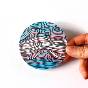 May include: A round sticker with a wavy pattern in blue, pink, and white colors. The waves are outlined in black.