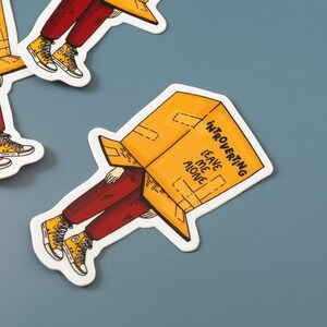 May include: A yellow and white sticker with a cartoon illustration of a person wearing a box on their head. The box has the text "Introverting Leave Me Alone" written on it.
