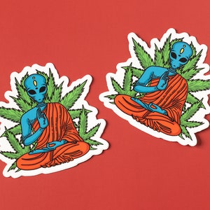 May include: Two blue alien figures in orange robes meditating in a lotus position surrounded by green marijuana leaves. The aliens have large eyes and are sitting on a white background.