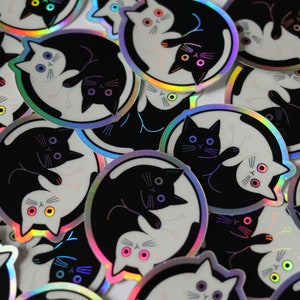 May include: A collection of iridescent stickers featuring black and white cats in a yin yang design. The stickers have a holographic finish and are perfect for adding a touch of whimsy to laptops, water bottles, or journals.