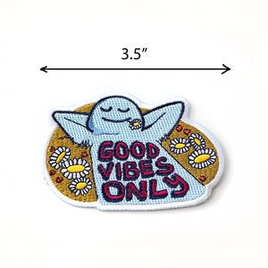 K&ouml;nnte beinhalten: Gestickter B&uuml;gel-Patch mit einer blauen Comicfigur mit dem Text "Good Vibes Only" umgeben von gelben G&auml;nsebl&uuml;mchen. Der Patch hat einen Durchmesser von 8,9 cm.