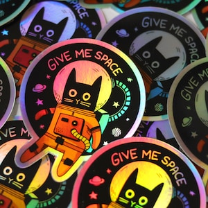 May include: A set of holographic stickers featuring a black cat wearing a space suit with the text "Give Me Space". The stickers are round and have a rainbow iridescent finish.