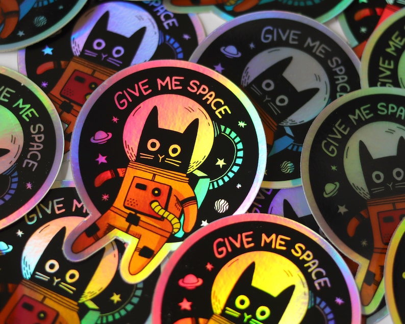 May include: A set of holographic stickers featuring a black cat wearing a space suit with the text "Give Me Space". The stickers are round and have a rainbow iridescent finish.
