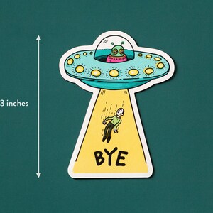 May include: A yellow and blue cartoon UFO with a green alien inside is beaming a person up with the word "BYE" written on the beam. The UFO is 3 inches tall.