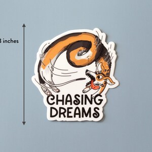 May include: A sticker with a cartoon corgi dog chasing its tail, with the text "CHASING DREAMS". The sticker is approximately 3 inches in height. The dog is colored in shades of brown, white, and black, with its tongue sticking out.