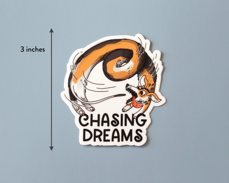 May include: A sticker with a cartoon corgi dog chasing its tail, with the text "CHASING DREAMS". The sticker is approximately 3 inches in height. The dog is colored in shades of brown, white, and black, with its tongue sticking out.