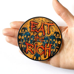 May include: A round sticker with a yellow background and black outline. The sticker features a stylized illustration of a crowd of faces with the text "Eat the Rich" in red and black lettering.