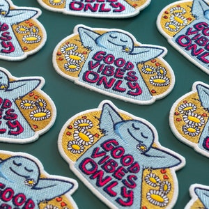 K&ouml;nnte beinhalten: Ein Set mit acht gestickten B&uuml;geletiketten mit einem Comic-Charakter und dem Text "Good Vibes Only" in rosa, blau und gelben Buchstaben. Die Patches sind von einem gelben Hintergrund mit wei&szlig;en G&auml;nsebl&uuml;mchen umgeben.
