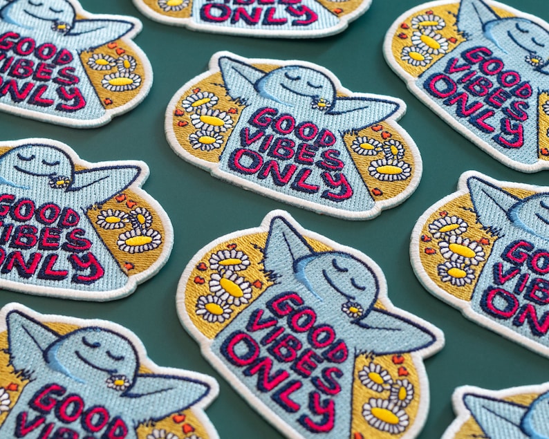 K&ouml;nnte beinhalten: Ein Set mit acht gestickten B&uuml;geletiketten mit einem Comic-Charakter und dem Text "Good Vibes Only" in rosa, blau und gelben Buchstaben. Die Patches sind von einem gelben Hintergrund mit wei&szlig;en G&auml;nsebl&uuml;mchen umgeben.