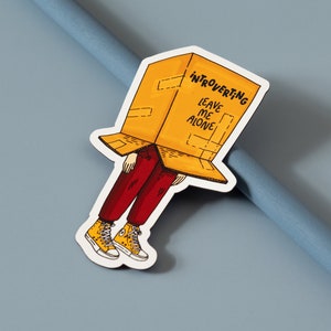 May include: A yellow cardboard box with the text "Introverting Leave Me Alone" on it. A person with red pants and yellow shoes is wearing the box as a hat.