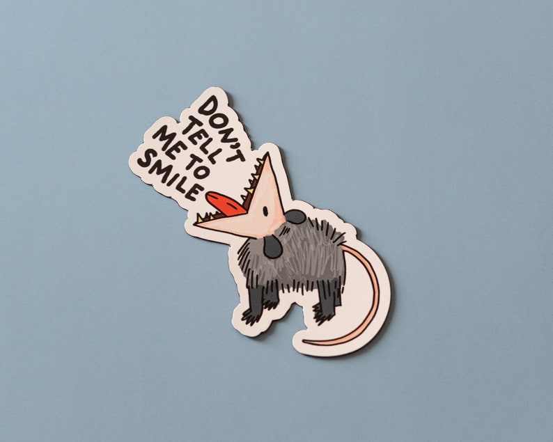 May include: A sticker featuring a cartoon opossum with its mouth open and the text "Don't tell me to smile".