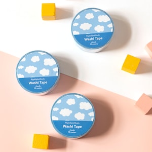 May include: Three rolls of washi tape with a blue sky and cloud pattern. Each roll has a blue label with the text "Washi Tape clouds 10 meters". The background includes pink, white, and yellow wooden blocks.
