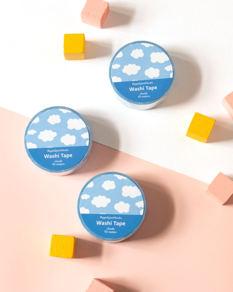 May include: Three rolls of washi tape with a blue sky and cloud pattern. Each roll has a blue label with the text "Washi Tape clouds 10 meters". The background includes pink, white, and yellow wooden blocks.