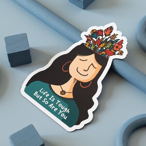 May include: A sticker featuring a cartoon illustration of a person with long black hair and flowers growing out of their head. The person is smiling and the text "Life is Tough But So Are You" is written on the sticker.
