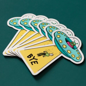 May include: A set of six yellow and blue stickers featuring a cartoon alien in a flying saucer. The stickers have the word "BYE" written on them.