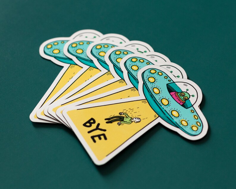 May include: A set of six yellow and blue stickers featuring a cartoon alien in a flying saucer. The stickers have the word "BYE" written on them.