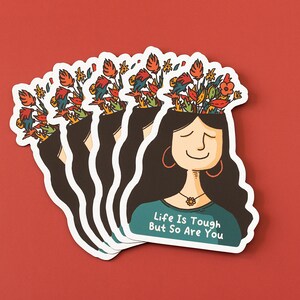 May include: A stack of stickers featuring a woman with flowers growing out of her head. The woman is smiling and has long dark hair. The text on the sticker reads "Life is tough but so are you."