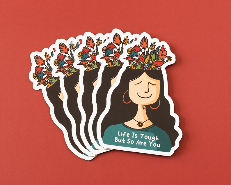 May include: A stack of stickers featuring a woman with flowers growing out of her head. The woman is smiling and has long dark hair. The text on the sticker reads "Life is tough but so are you."