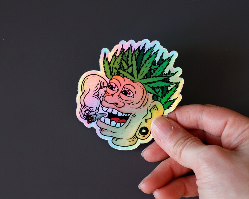 Peut inclure: Autocollant holographique repr&eacute;sentant un visage de dessin anim&eacute; avec des feuilles de cannabis vertes en guise de cheveux, un joint allum&eacute; et un large sourire. L'autocollant a un &eacute;clat arc-en-ciel et une boucle d'oreille noire.