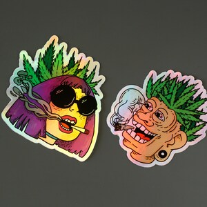Peut inclure: Deux autocollants holographiques avec des illustrations de dessins anim&eacute;s. L'un montre une personne aux cheveux violets, des lunettes de soleil et une couronne de feuilles de cannabis, exhalant de la fum&eacute;e. L'autre repr&eacute;sente une personne aux cheveux de cannabis verts, fumant un joint.