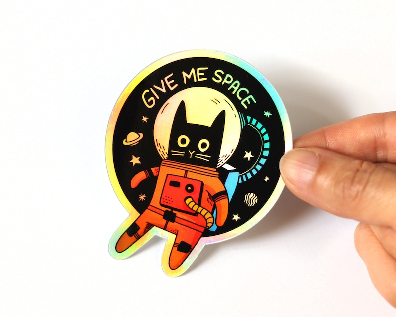May include: A holographic sticker featuring a black cat wearing a red space suit with the text "Give Me Space" in a rainbow font.