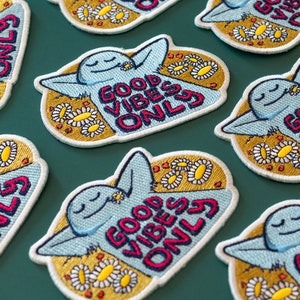 K&ouml;nnte beinhalten: Ein Set gestickter B&uuml;gelbilder mit einer blauen Comicfigur mit dem Text "Good Vibes Only" in rot, wei&szlig; und blauen Buchstaben. Die Patches sind von gelben G&auml;nsebl&uuml;mchen umgeben.