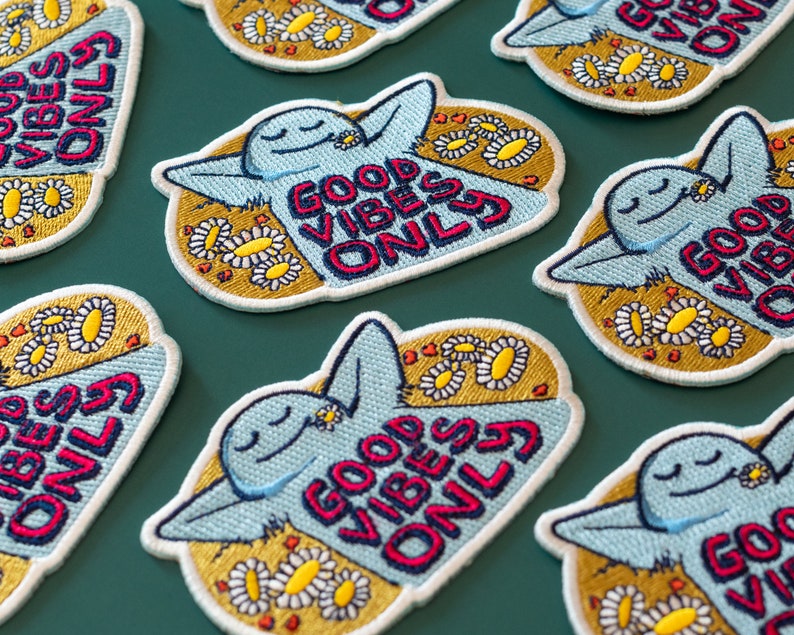 K&ouml;nnte beinhalten: Ein Set gestickter B&uuml;gelbilder mit einer blauen Comicfigur mit dem Text "Good Vibes Only" in rot, wei&szlig; und blauen Buchstaben. Die Patches sind von gelben G&auml;nsebl&uuml;mchen umgeben.