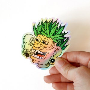 Peut inclure: Autocollant holographique repr&eacute;sentant un visage de dessin anim&eacute; avec des cheveux en forme de feuilles de cannabis vertes, un joint allum&eacute; et un large sourire. L'autocollant a une finition iris&eacute;e et une boucle d'oreille noire. L'image est tenue par une main.