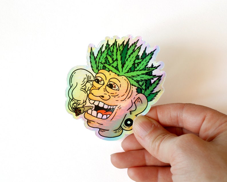 Peut inclure: Autocollant holographique repr&eacute;sentant un visage de dessin anim&eacute; avec des cheveux en forme de feuilles de cannabis vertes, un joint allum&eacute; et un large sourire. L'autocollant a une finition iris&eacute;e et une boucle d'oreille noire. L'image est tenue par une main.