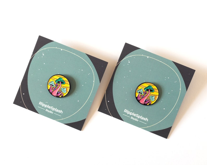 May include: Two enamel pins featuring a colorful mushroom design with a yellow and orange background. The pins are on a black and green background with the text "RippleSplash Studio" on each card.