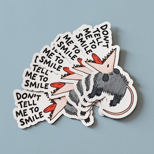 May include: A set of six stickers featuring a cartoon opossum with the text "Don't tell me to smile". The opossum is gray with a pink mouth and red tongue. The stickers are white with black text.