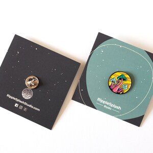 May include: A colorful enamel pin featuring a cluster of pink and green mushrooms with yellow accents. The pin is on a black card with the text "RippleSplash Studio" and a website address.