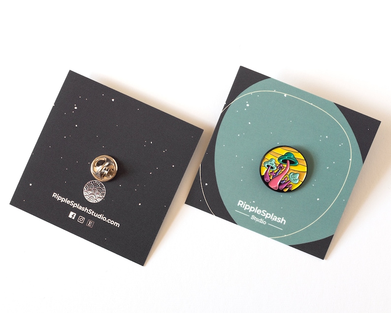 May include: A colorful enamel pin featuring a cluster of pink and green mushrooms with yellow accents. The pin is on a black card with the text "RippleSplash Studio" and a website address.