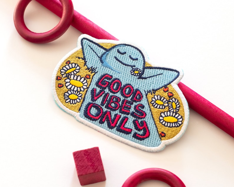 K&ouml;nnte beinhalten: Ein blauer gestickter Patch mit einer Comicfigur mit geschlossenen Augen und einer G&auml;nsebl&uuml;mchen in den Haaren. Der Patch hat gelbe G&auml;nsebl&uuml;mchen und den Text "GOOD VIBES ONLY" in Rot und Blau.