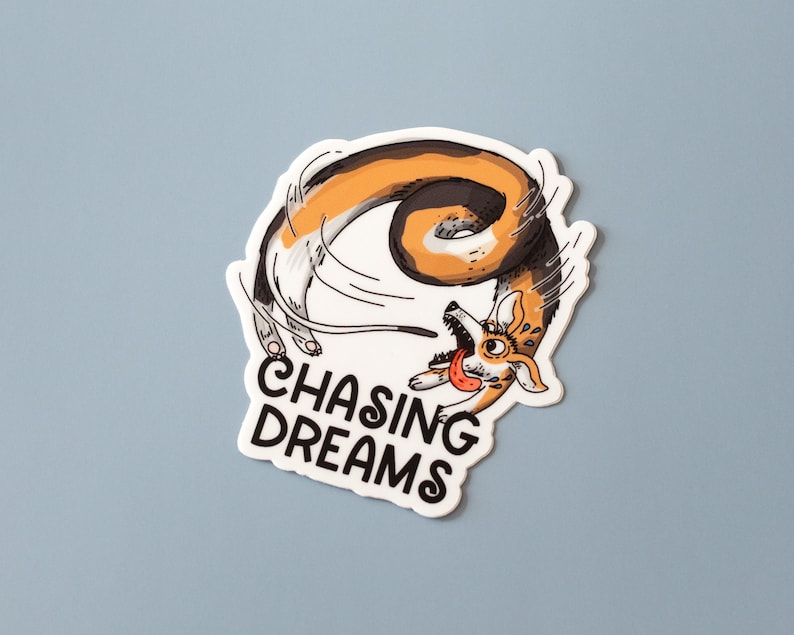 May include: A sticker featuring a cartoon corgi chasing its tail. The corgi is orange, white, and brown, with its tongue sticking out. The sticker has the words "CHASING DREAMS" in a bold, black font.