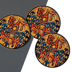 May include: Three round stickers with an orange background and black outlines. Each sticker features a stylized illustration of a crowd of faces with the text "Eat the Rich" in red and blue.