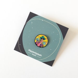 May include: A colorful enamel pin featuring a cluster of pink and purple mushrooms with green tops, set against a yellow and orange background. The pin is on a card with the text "RippleSplash Studio".